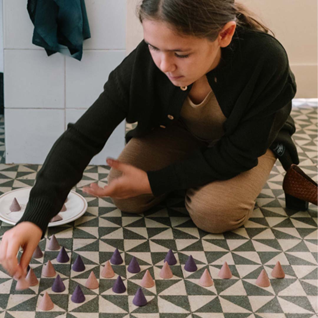Een kind knielt op de grond en rangschikt grapat houten kegels in paarse en beige kleuren.
