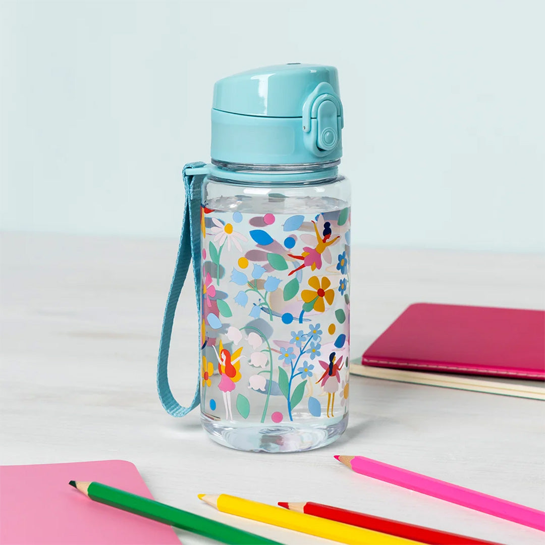 Een Rex London drinkfles met feeën en bloemen, gevuld met water, staat op een bureau met kleurpotloden en notitieboekjes.