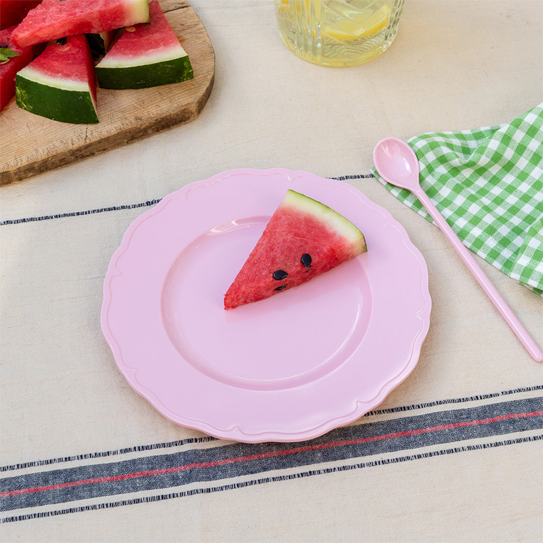 Een Rex London roze melamine kinderbord met een stuk watermeloen erop, op een gestreepte tafelkleed.