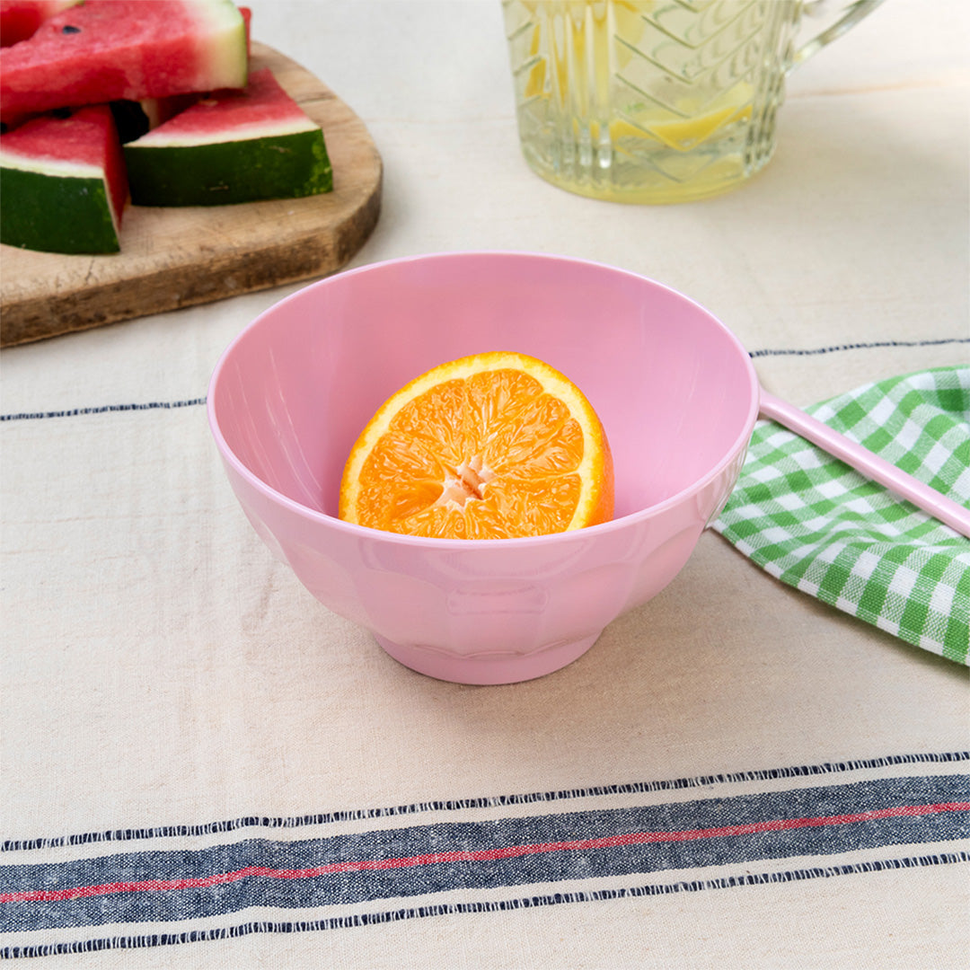 Een roze rex london melamine kom met een halve sinaasappel op een gestreepte tafelkleed.