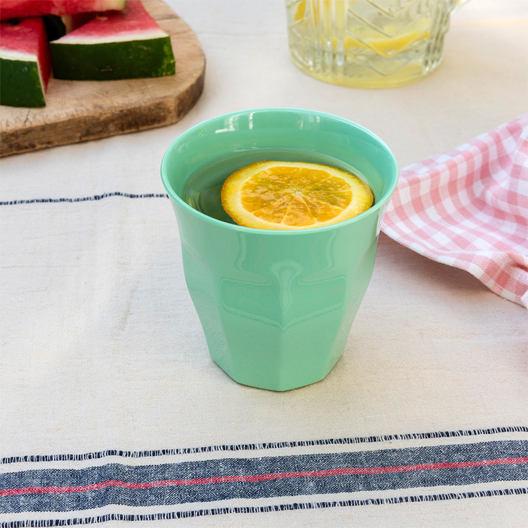 Een pistachegroene kinderbeker met water en een sinaasappelschijfje op een buitentafel.