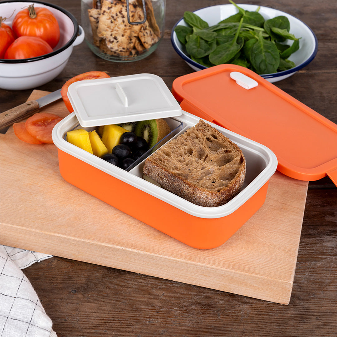 Rex London oranje bento-broodtrommel gevuld met brood en fruit op een houten snijplank.