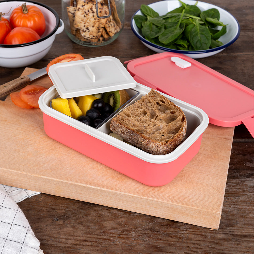 Open roze broodtrommel met een boterham en fruit op een houten snijplank.