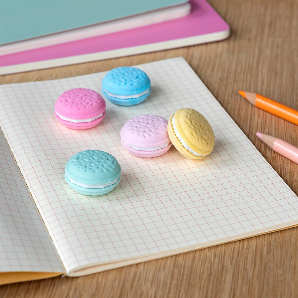 Vijf kleurrijke macaron gummen liggen op een open schrift met ruitjespapier, met kleurpotloden op de achtergrond.