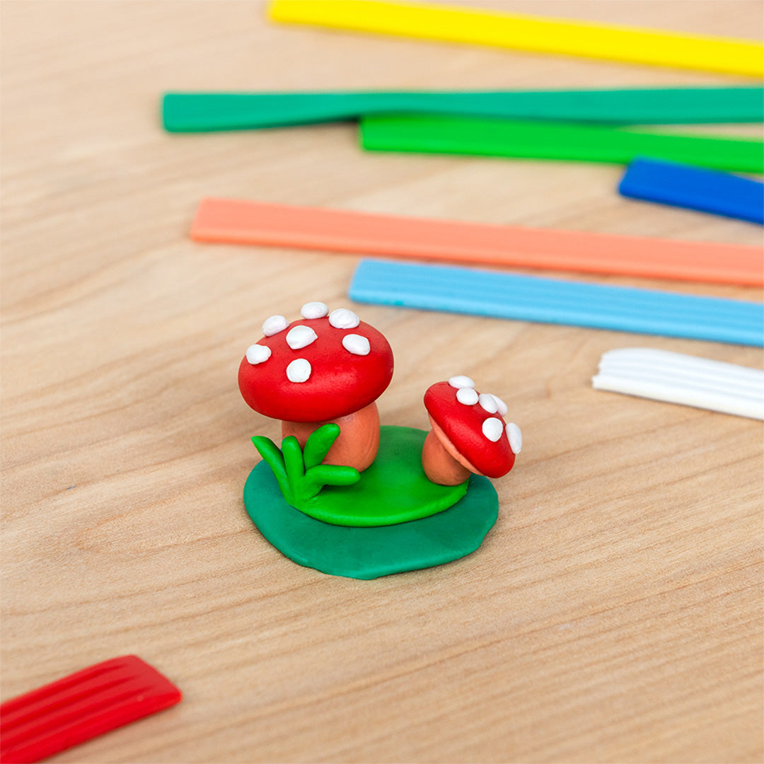 Twee boetseerklei paddestoelen op een groene ondergrond, met gekleurde kleisticks op een houten tafel.