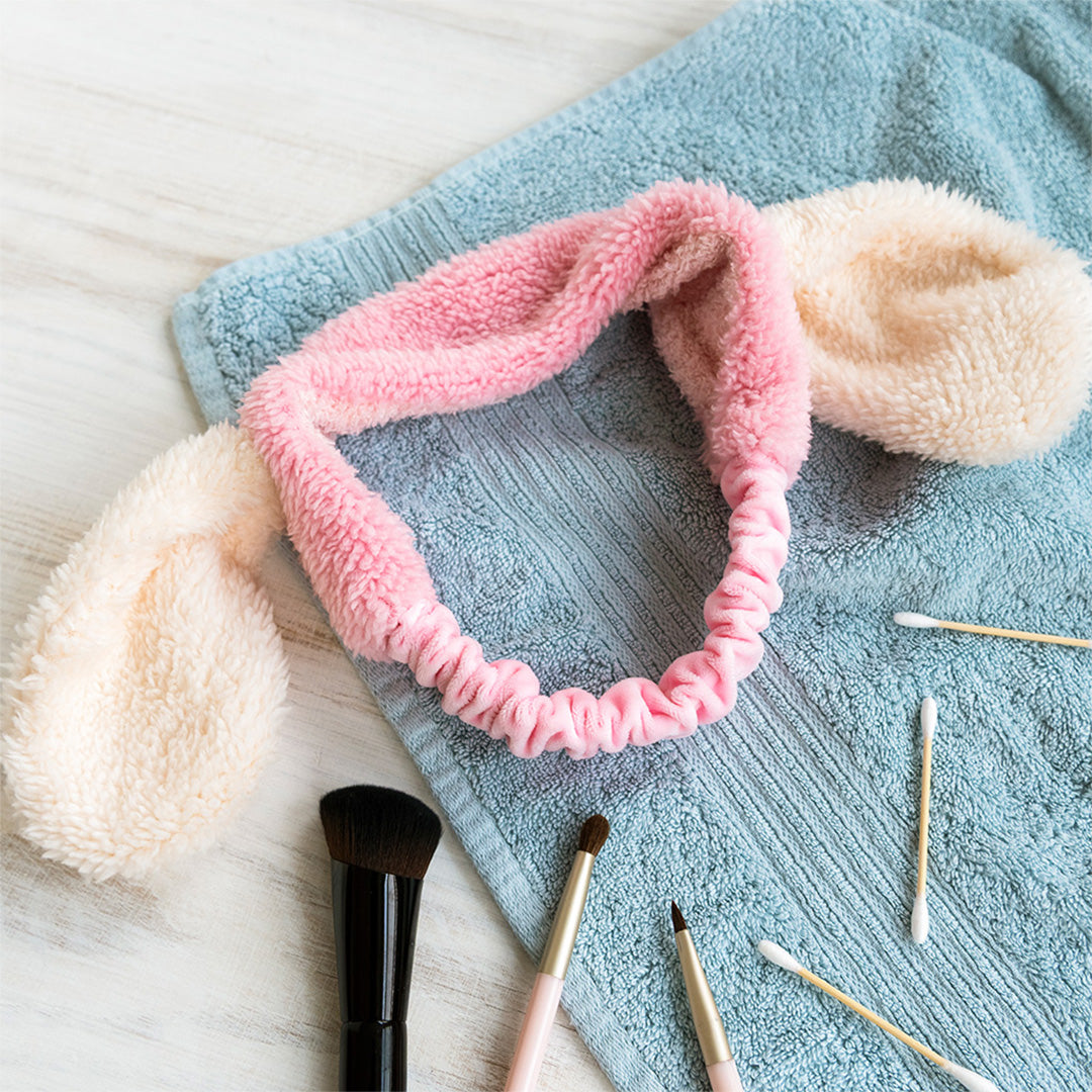 Rex London haarband met roze en crèmekleurige pluizige bunny ears op een blauwe handdoek met make-upkwasten.