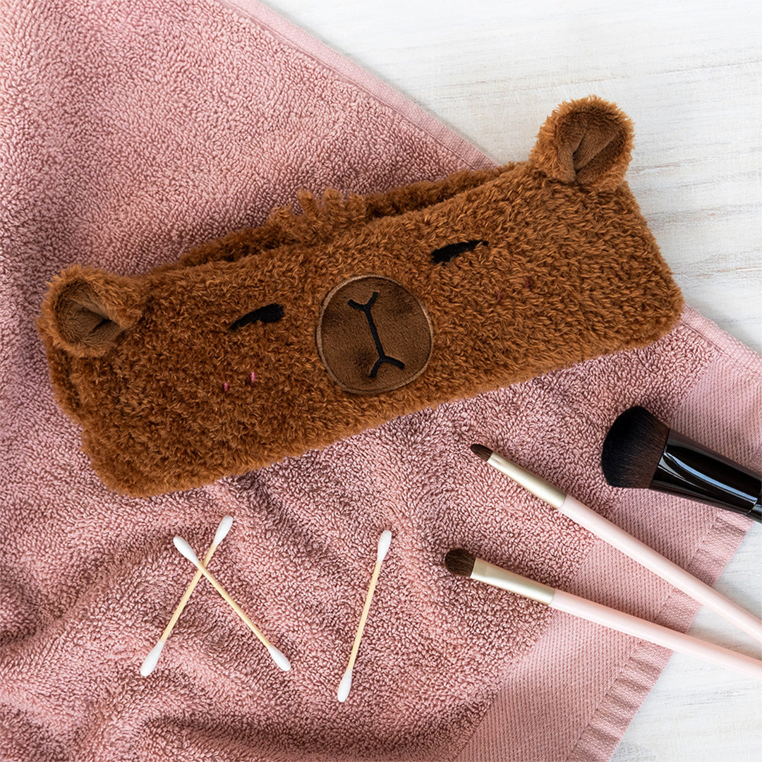 De bruine capybara haarband ligt op een roze handdoek, met make-upkwasten en wattenstaafjes ernaast.