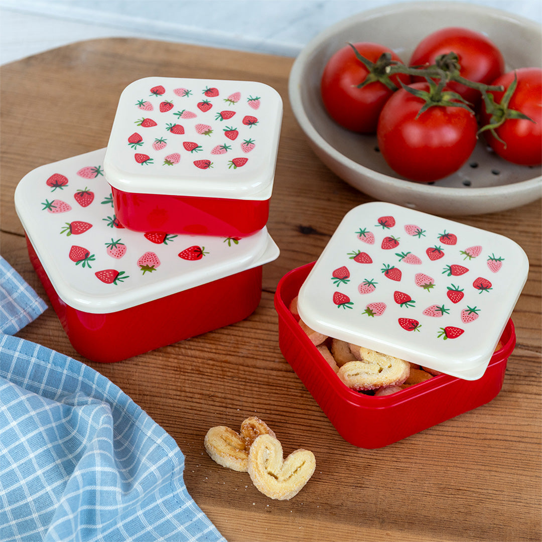 Drie Rex London snackdoosjes met aardbeipatroon op een houten ondergrond, waarvan één open met koekjes.