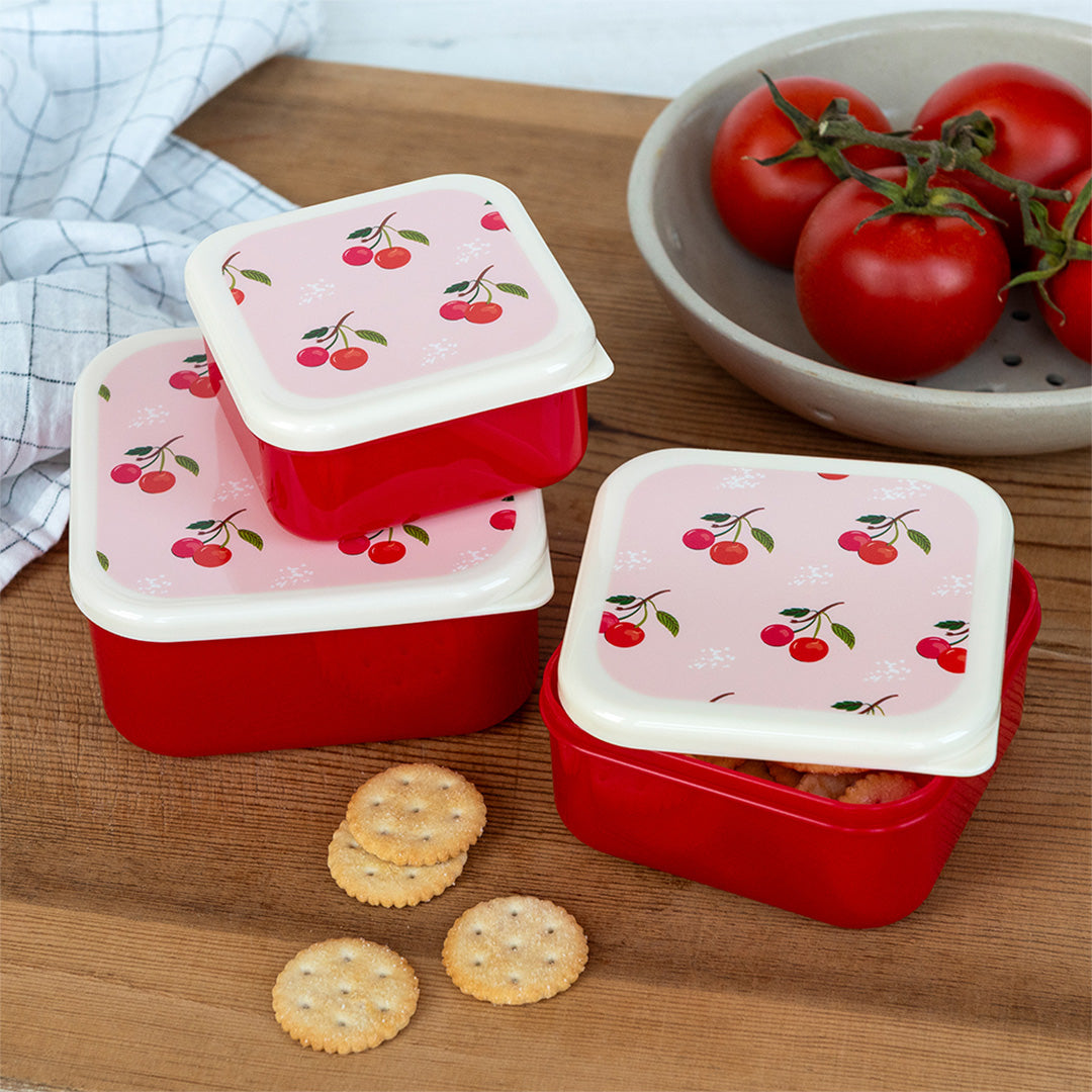 Drie rode Rex London snackdoosjes met kersenpatroon op een houten ondergrond, waarvan één open met crackers.