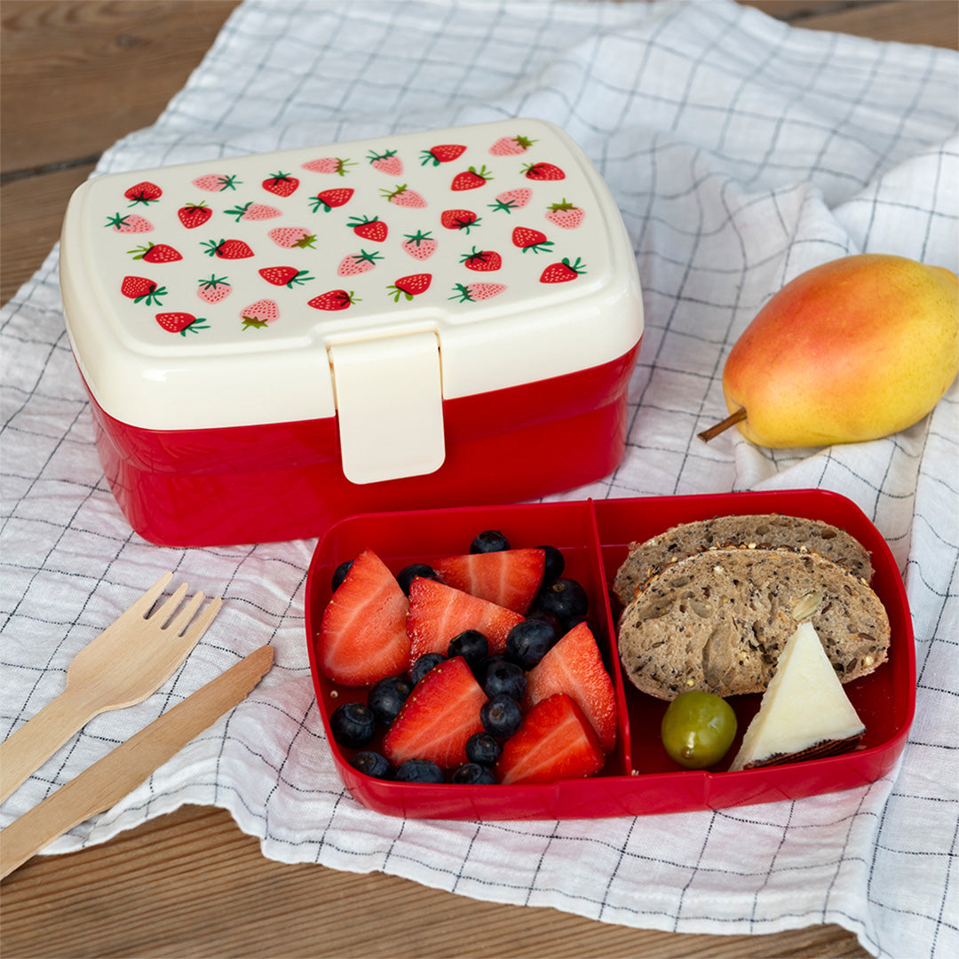 Open Rex London broodtrommel met aardbeienprint, gevuld met fruit en brood, op een geruite doek naast een peer.