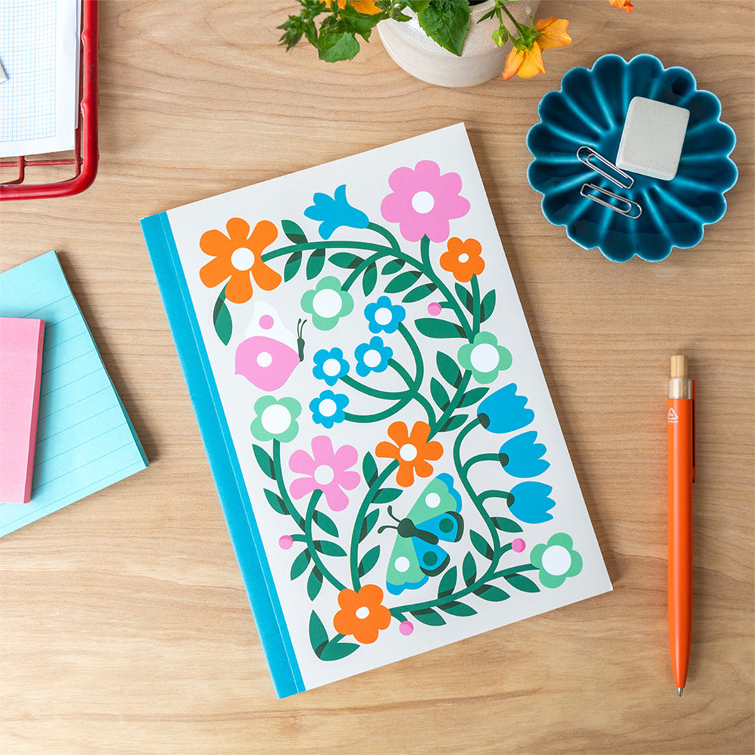 Rex London A5 schrift met bloemen- en vlinderpatroon liggend op een houten bureau met kantoorartikelen.