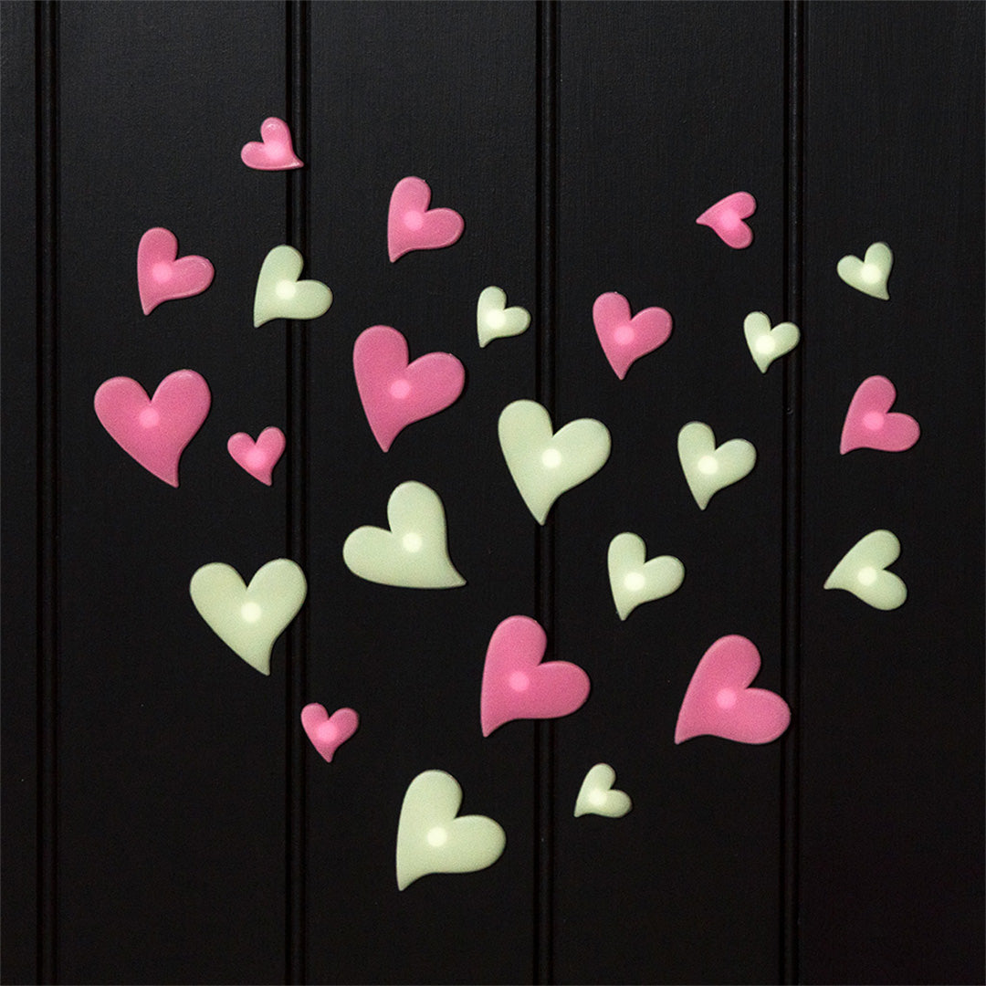 Een verzameling roze en lichtgroene glow-in-the-dark hartjes decoratiestickers op een zwarte ondergrond.