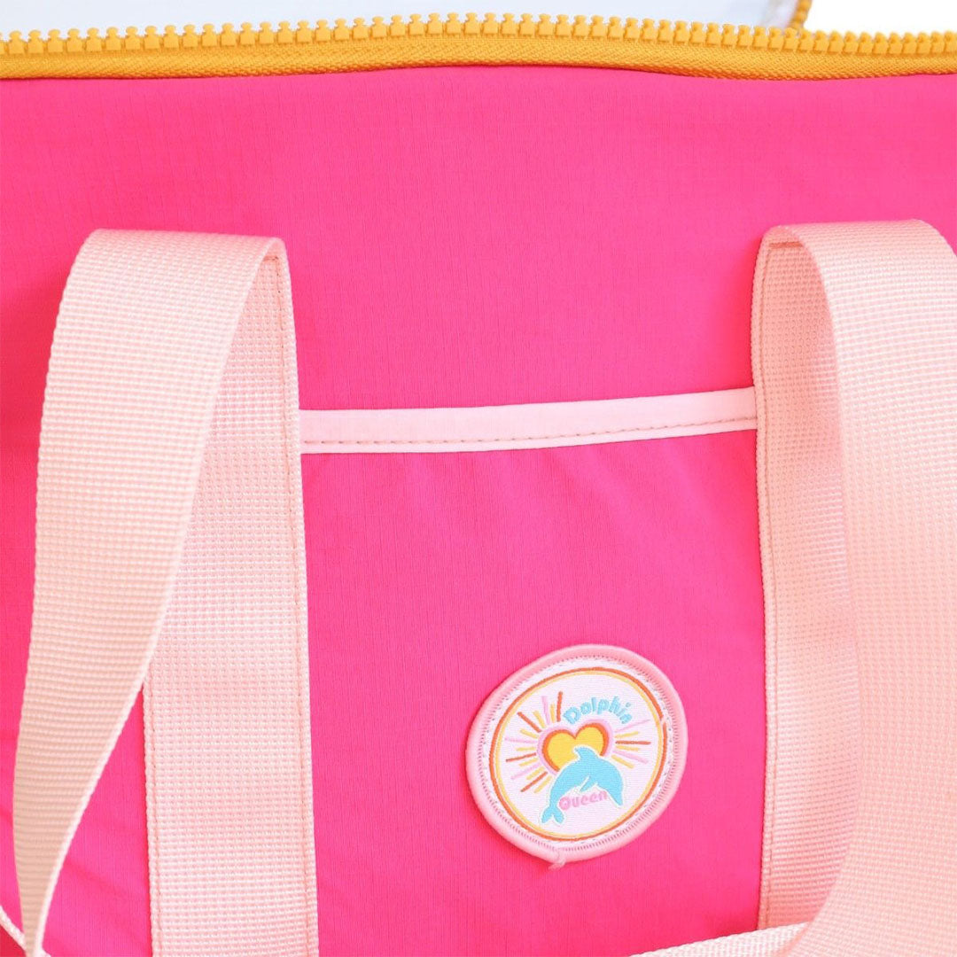 Close-up van de le petit souk koeltas met een rond dolfijnlogo en de tekst 'Dolphin Queen' op de roze stof.