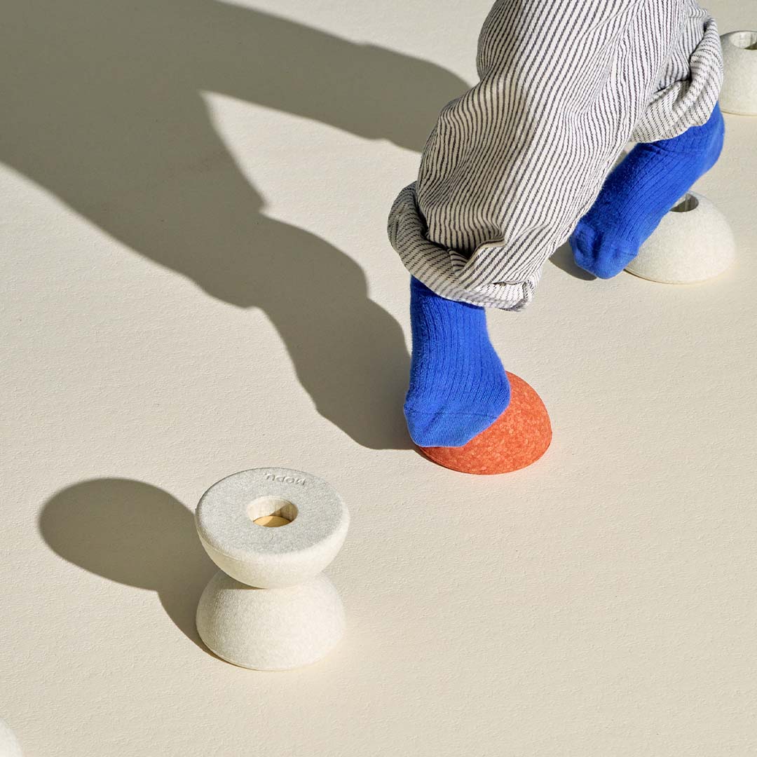 Kind met blauwe sokken stapt op een oranje modu schuimrubberen bal, met een zandgrijze bal eronder als onderdeel van bouwspeelgoed.