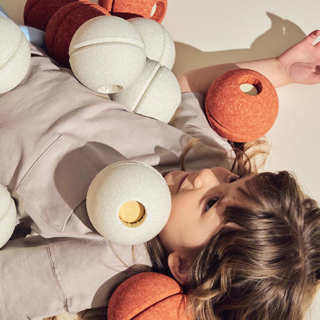 Kind ligt op de grond omringd door een collectie modu schuimrubberen ballen in zandgrijs en oranje.
