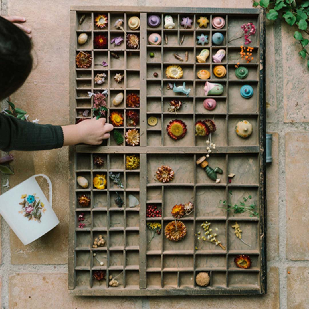 Een kinderhand pakt een van de Grapat houten speelstukken uit een grote houten sorteerbak gevuld met gedroogde bloemen en andere natuurlijke materialen.