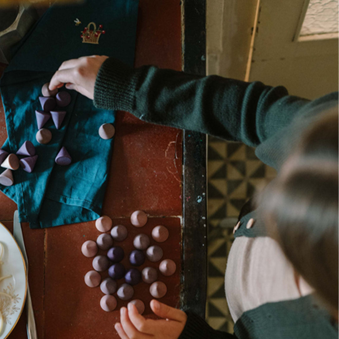 Bovenaanzicht van kinderhanden die grapat houten kegels in paarse en beige tinten rangschikken op een vloer.