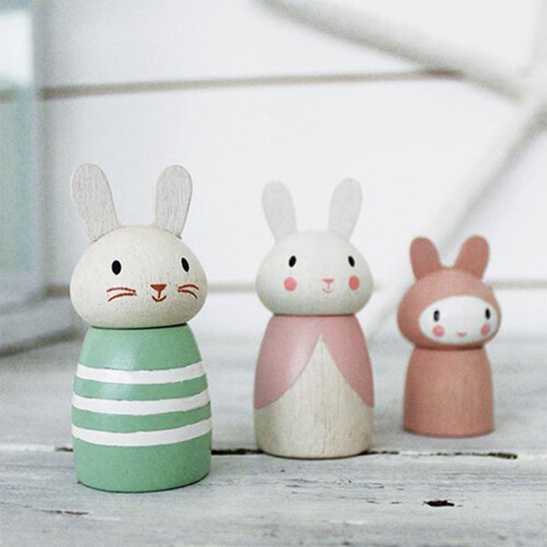 Drie Tender Leaf Toys houten konijnenfiguren, waarvan één groen gestreept, één roze met jurk en één kleine roze, staan op een lichte ondergrond.