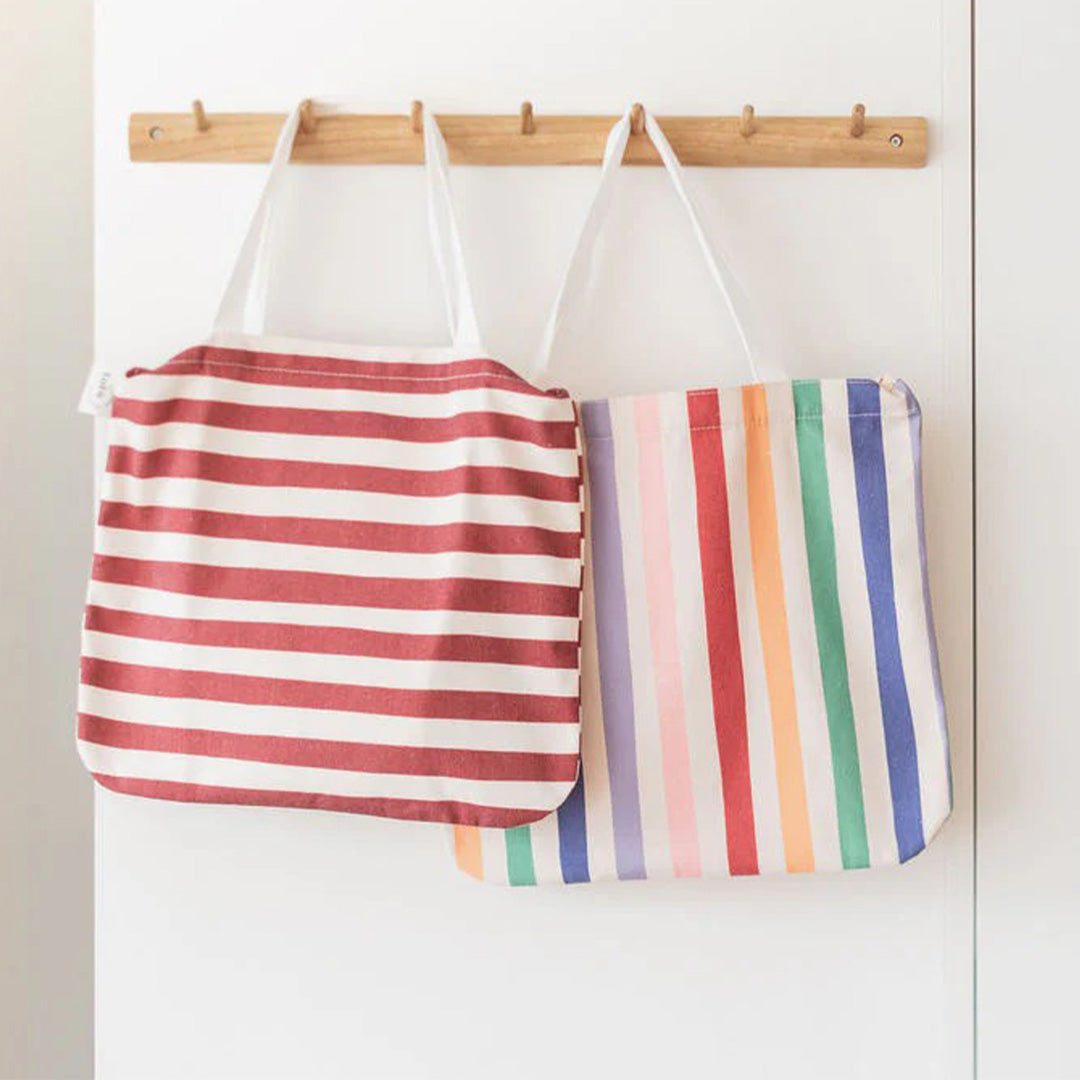 Twee play&go draagtassen met strepen, één rood-wit en één regenboogkleurig, hangen aan een houten kapstok.