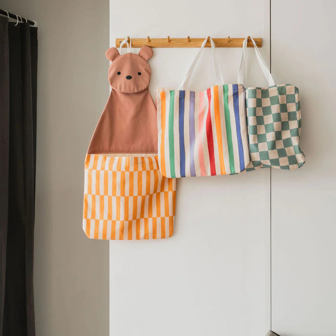 Een play&go wandopberger in de vorm van een beer en twee draagtassen met strepen en ruiten hangen aan een houten rek.