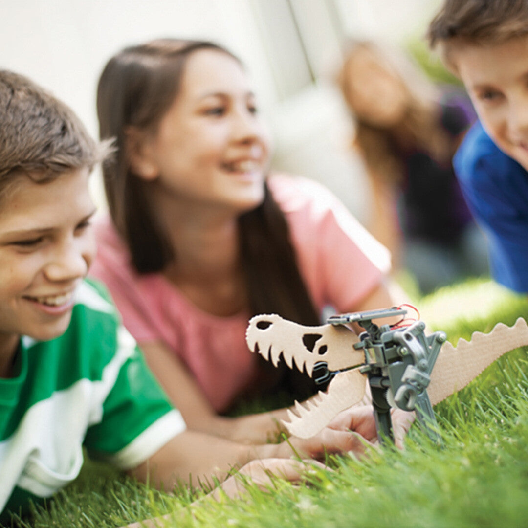 Twee kinderen liggen in het gras en kijken naar de 4M Solar Robot in dinosaurusvorm die voor hen staat.
