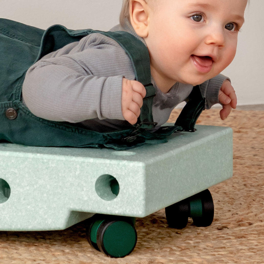 Een baby ligt op de buik op een lichtgroen modu rijdend speelgoed met groene wielen.
