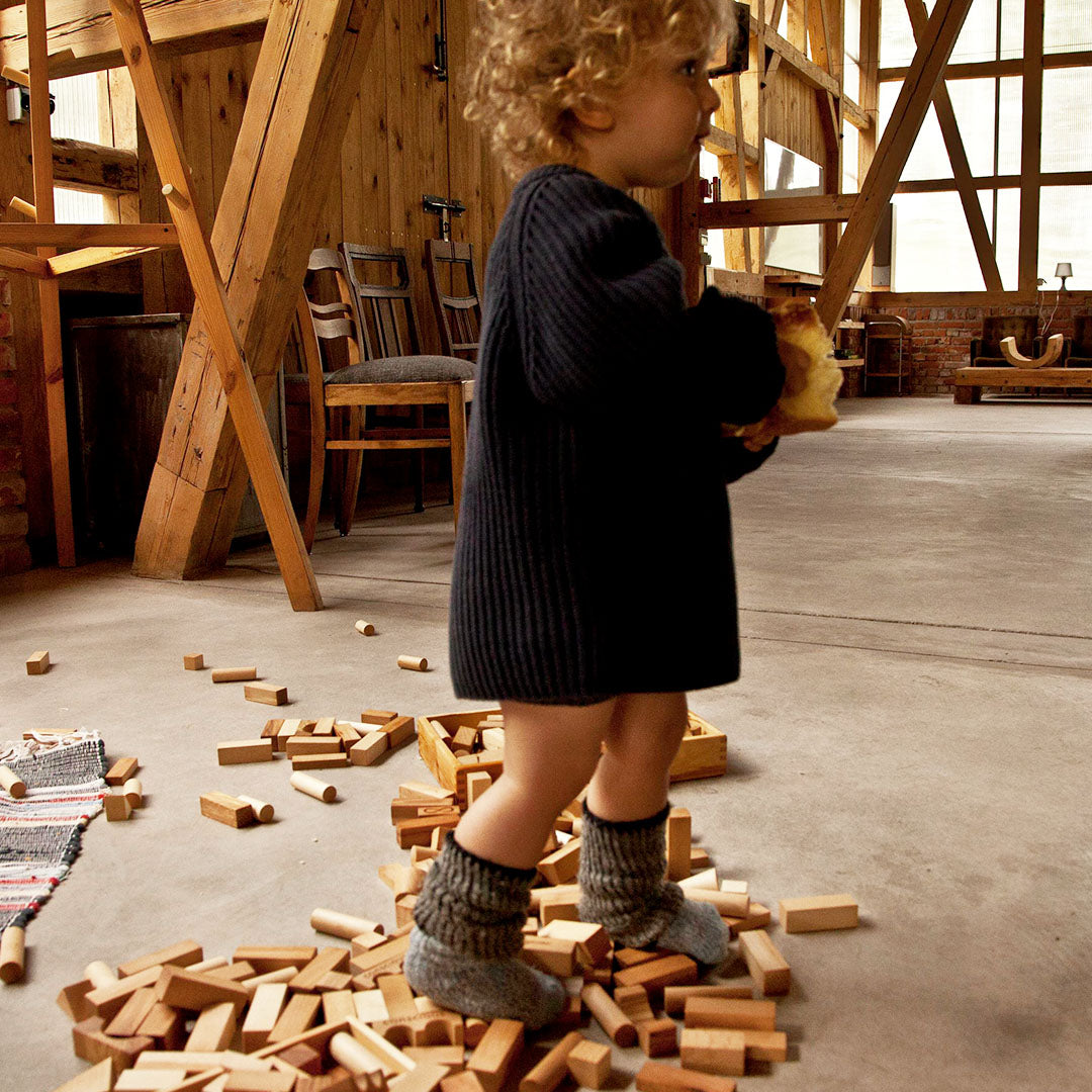 Een klein kind staat op een grote stapel Wooden Story houten bouwblokken op de grond.