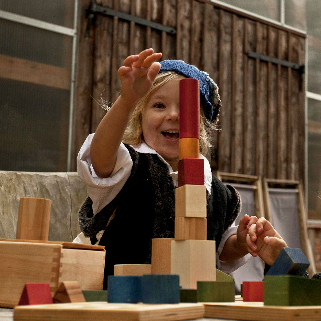 Een lachend kind speelt buiten met Wooden Story houten bouwblokken en bouwt een hoge toren.