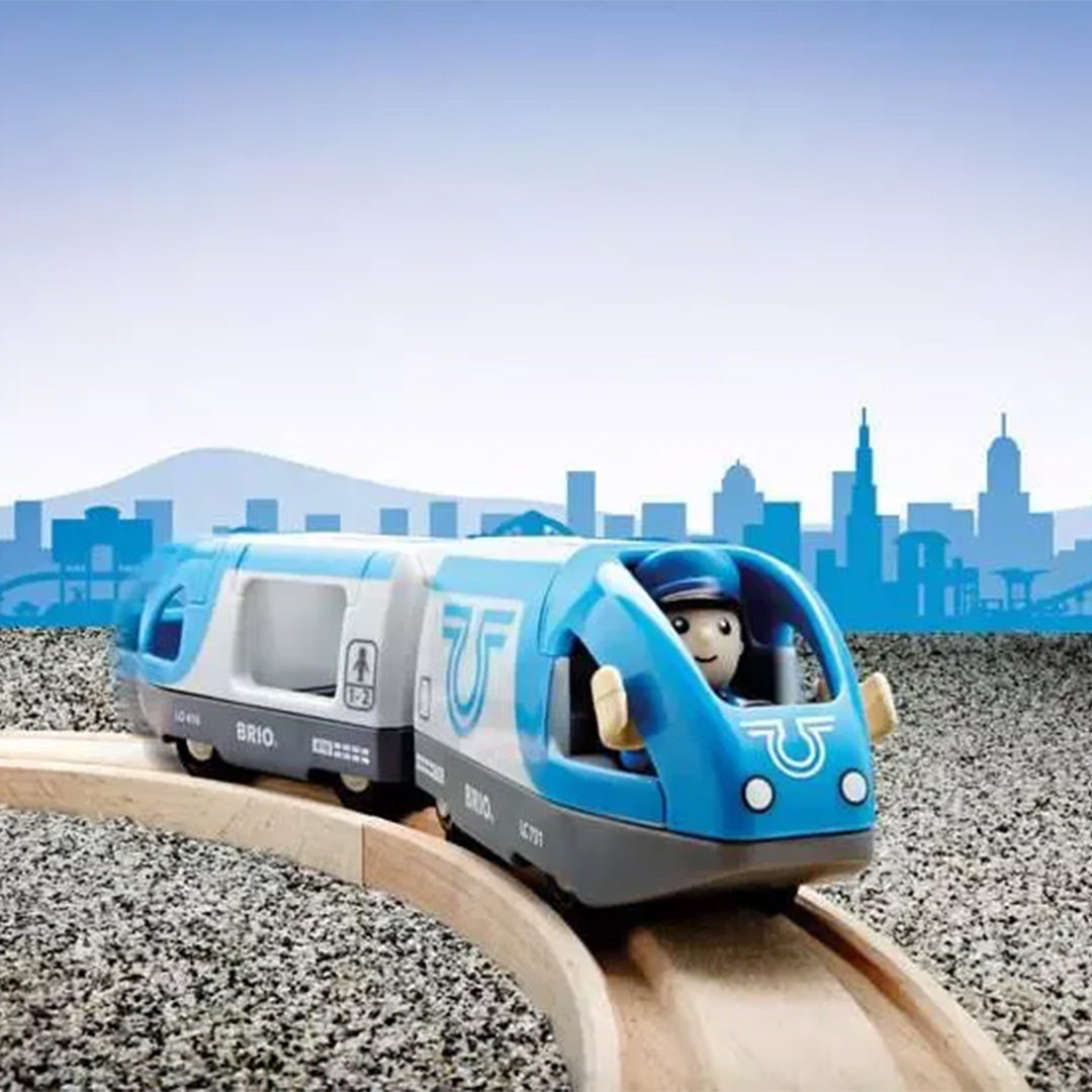 Brio passagierstrein speelgoed rijdend op houten rails met een conducteurfiguur in de blauw-witte trein, tegen een skyline achtergrond.