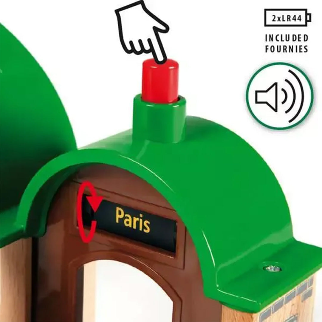 Detail van een Brio treinstation met een rode knop op het dak en een bordje 'Paris', met een hand die de knop aanwijst.