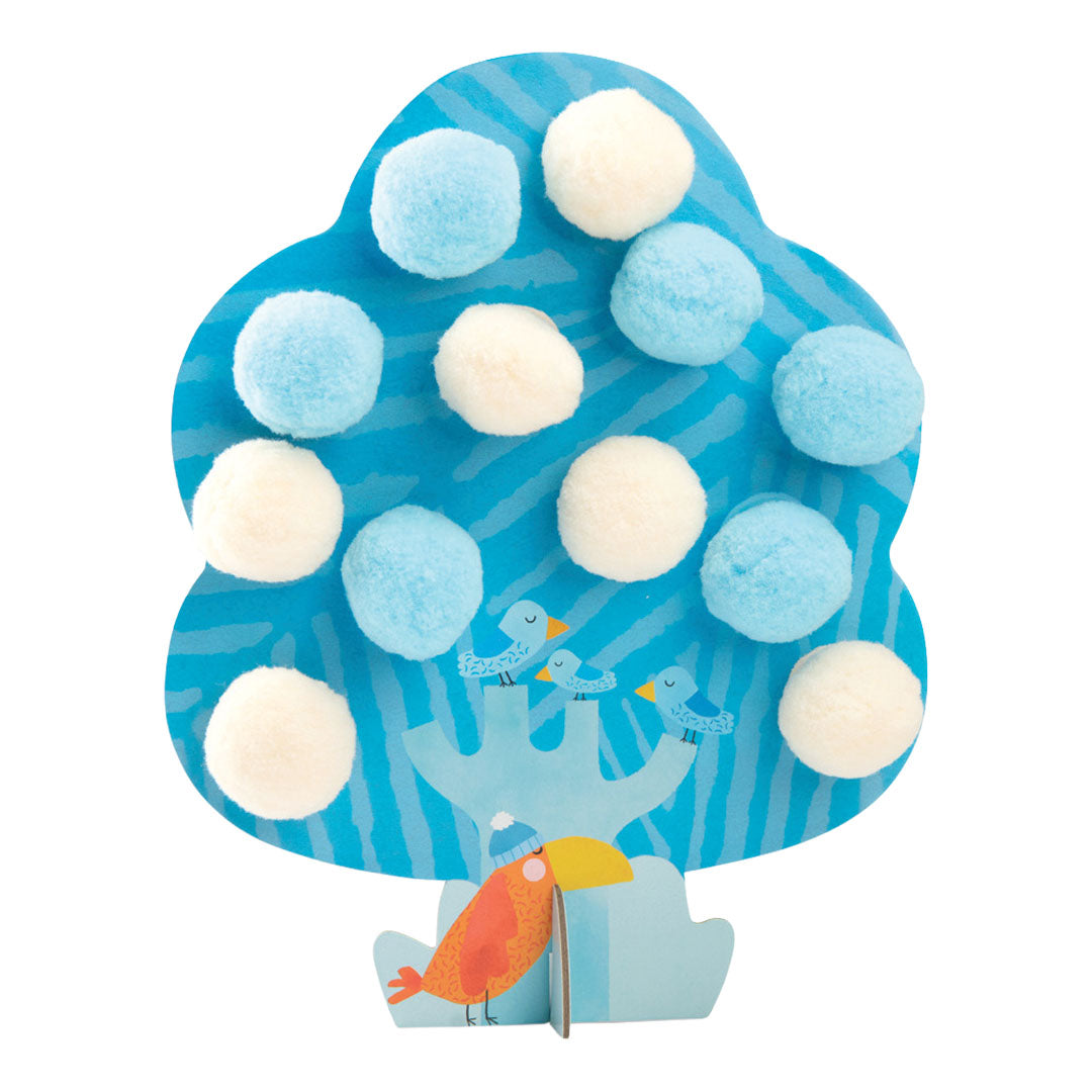Een blauwe kartonnen boom van de knutselset met lichtblauwe en witte pompoms en een vogelillustratie.