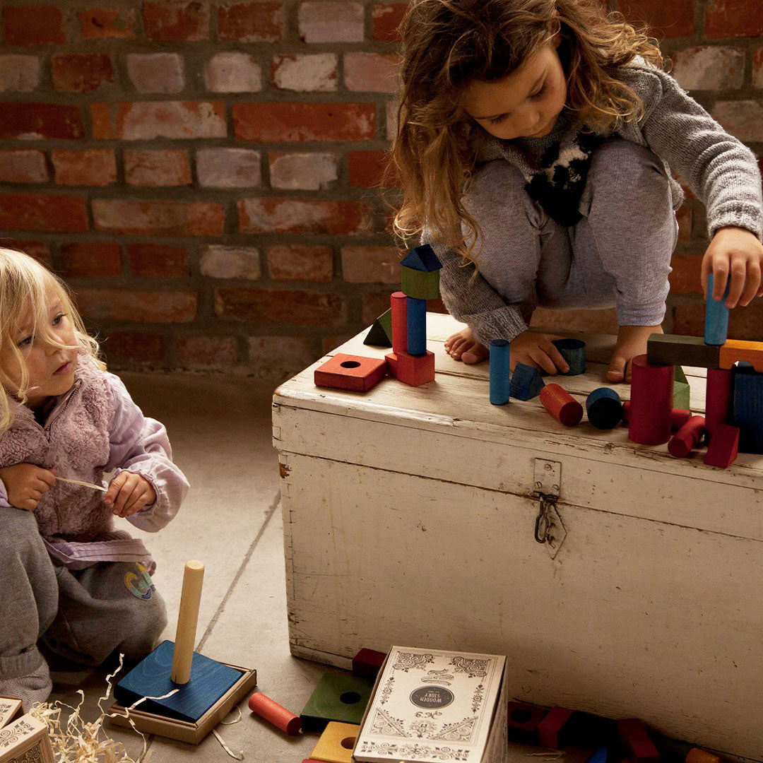 Twee kinderen spelen geconcentreerd met Wooden Story houten bouwblokken op en rond een witte kist.