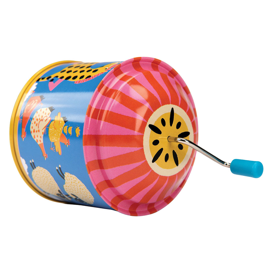 Moulin Roty muziekdoos van tin, liggend op de zijkant, met boerderijdieren en een gestreepte roze-rode draaihendel.