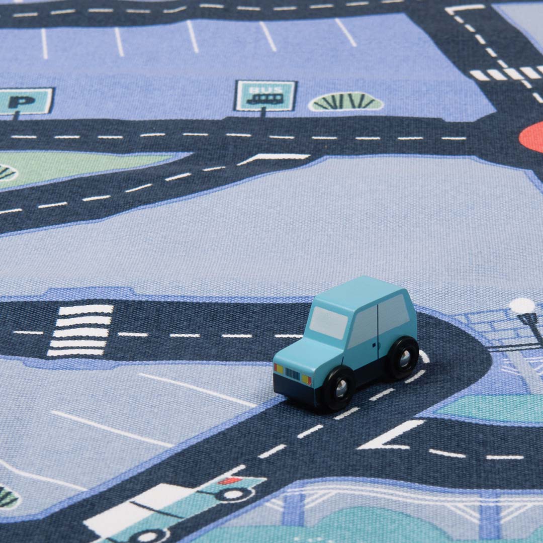 Detail van het Moulin Roty speelkleed met een lichtblauwe houten auto op een weg naast een parkeerplaats.