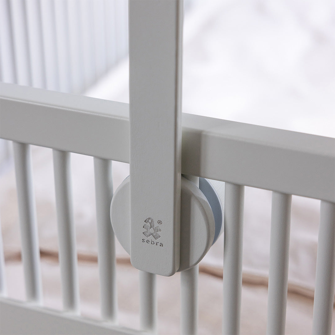 Detail van de Sebra witte houten mobielhouder met logo, bevestigd aan de spijlen van een babywieg.