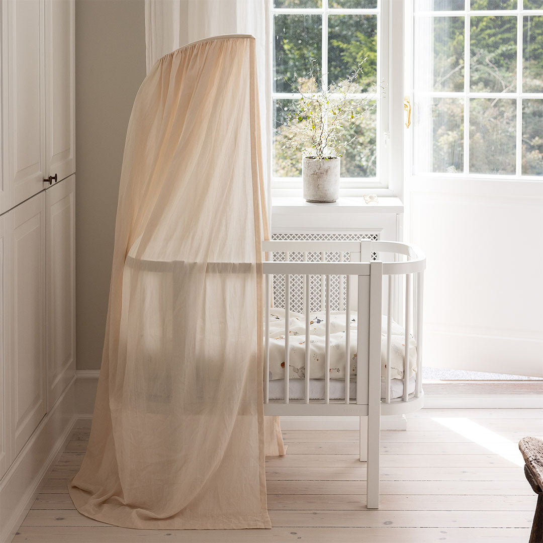 Witte babywieg met een lichtbeige hemeltje en de Sebra mobielhouder in een lichte kamer met raam.