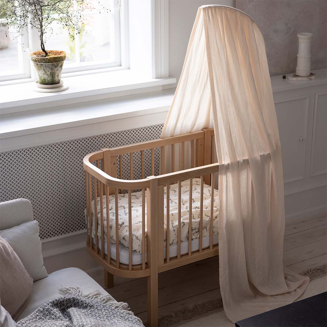 Een houten babywieg met een lichte stoffen hemel en beddengoed in een kamer.