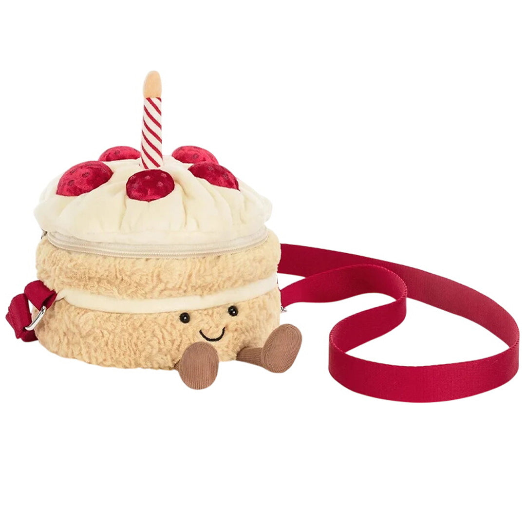 Jellycat kinder crossbodytas in de vorm van een verjaardagstaart met kaars en rode band, schuin gefotografeerd.