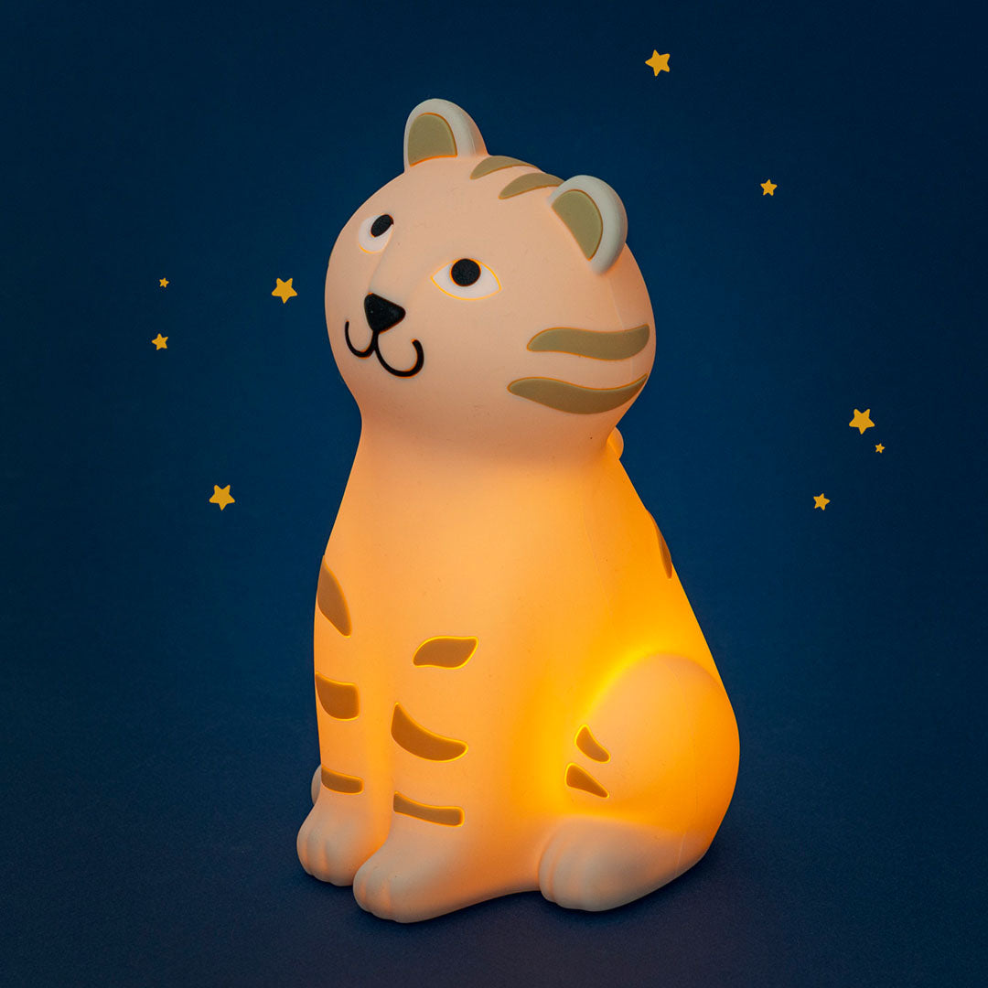 Moulin Roty nachtlampje in de vorm van een beige tijger dat warm wit licht geeft, schuin van voren gezien tegen een donkerblauwe achtergrond met sterren.