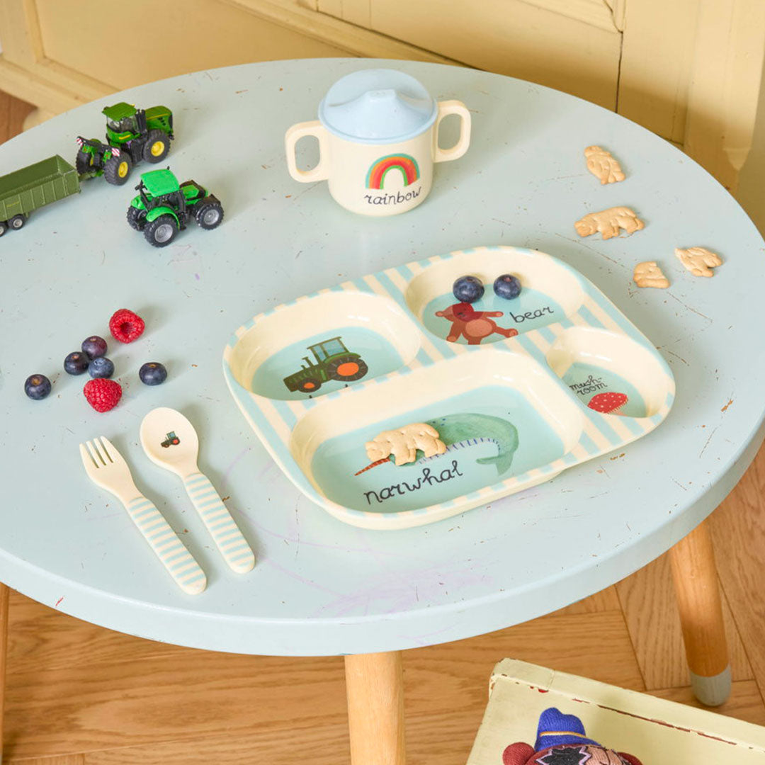 Een Rice kinderservies met een bord met tractor, beer en narwal, een regenboogbeker, lepel en vork op een blauwe tafel.