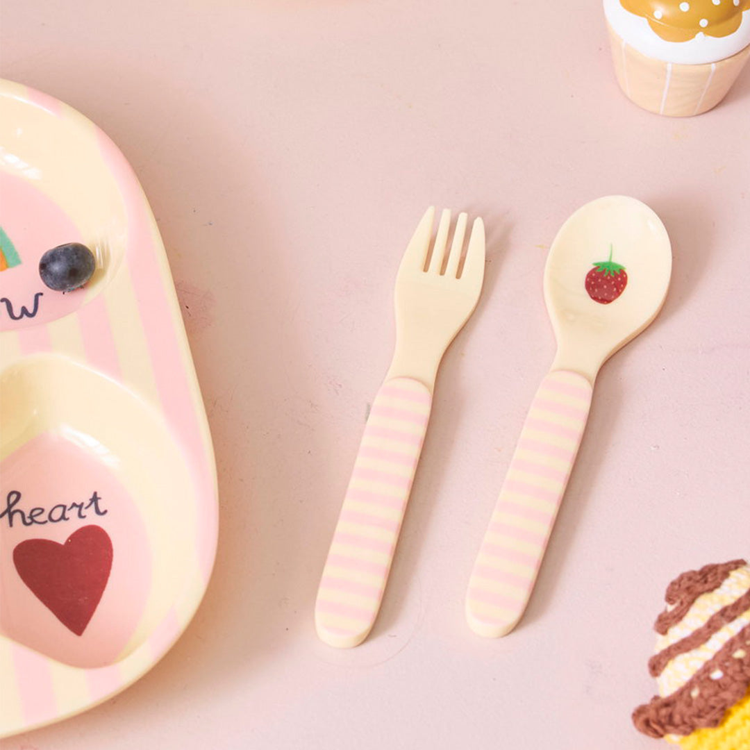 Kinderbestek set met lepel en vork met gestreepte handvatten, liggend op een roze ondergrond naast een bord.