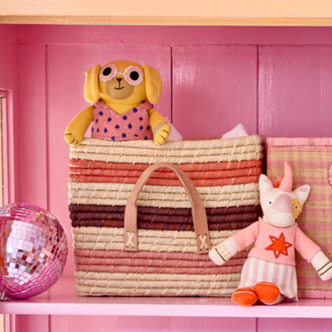 rice gestreepte raffia opbergmand op een roze plank, gevuld met een knuffelhond en een knuffeleenhoorn ernaast.