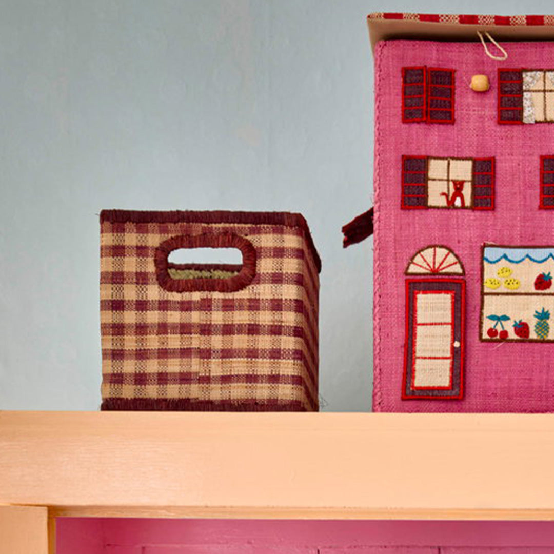 De rice raffia opbergmand met bruin geblokt patroon staat op een plank naast een roze speelgoedhuis.