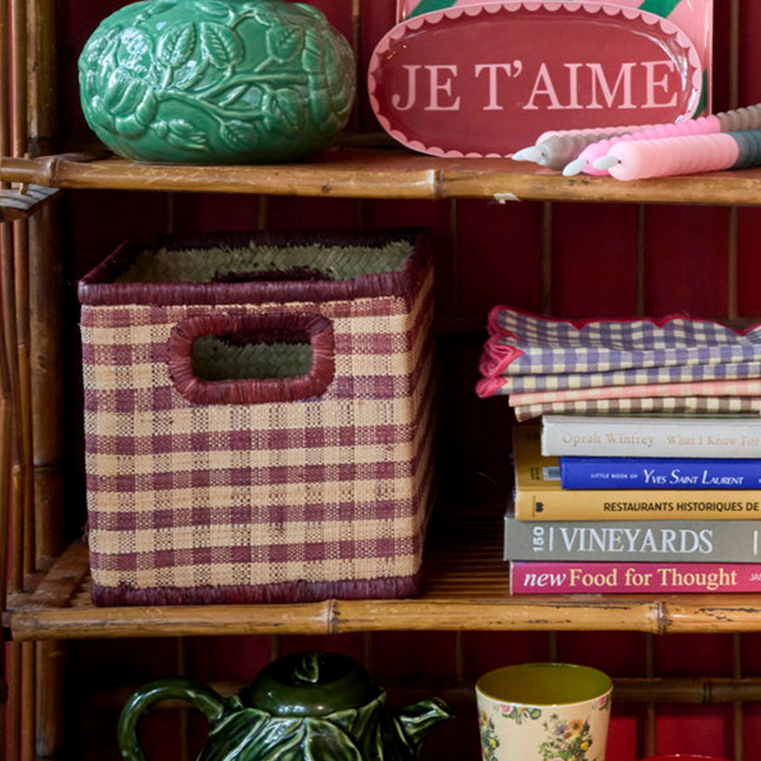 De rice raffia opbergmand met bruin geblokt patroon staat op een bamboe plank tussen boeken en decoratie.