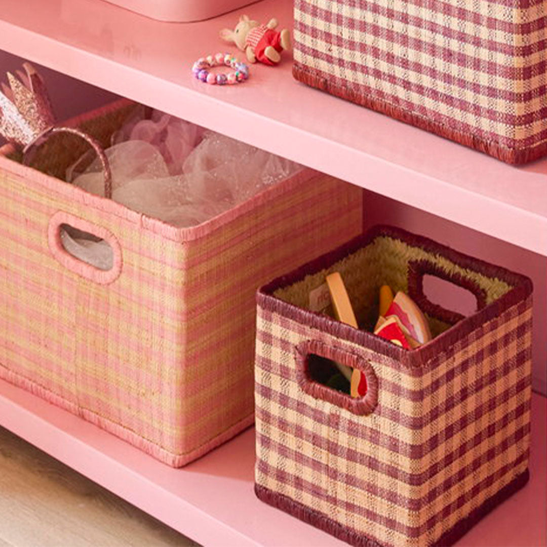 Twee rice raffia opbergmanden, één roze en één met bruin geblokt patroon, staan op een roze plank.