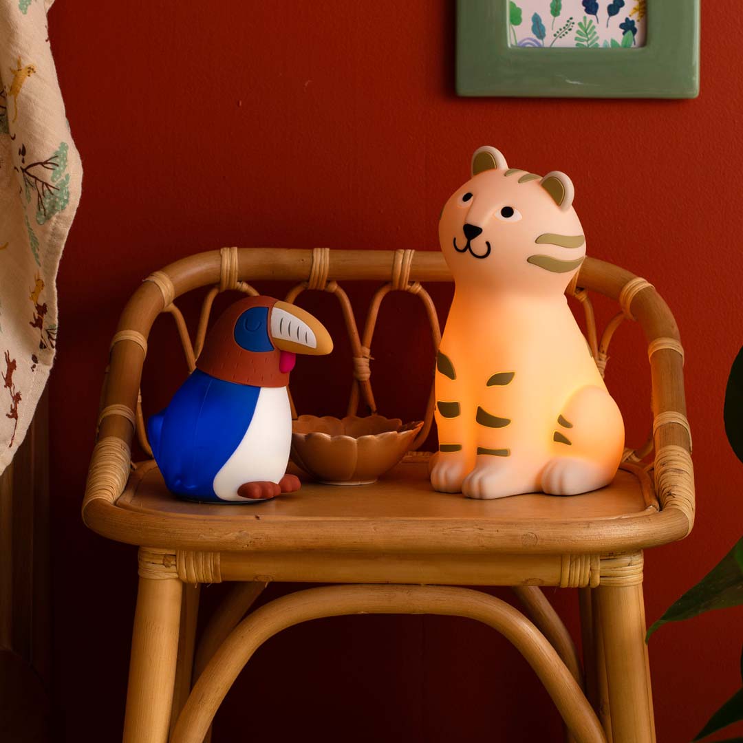 Moulin Roty nachtlampje in de vorm van een neushoornvogel staat naast een oplichtend tijger nachtlampje op een houten stoel.