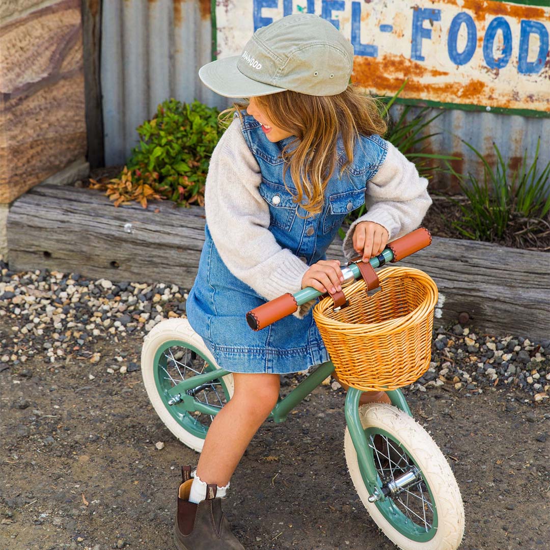 Een kind met een petje zit op de groene Banwood loopfiets met rieten mandje, kijkend naar de zijkant.