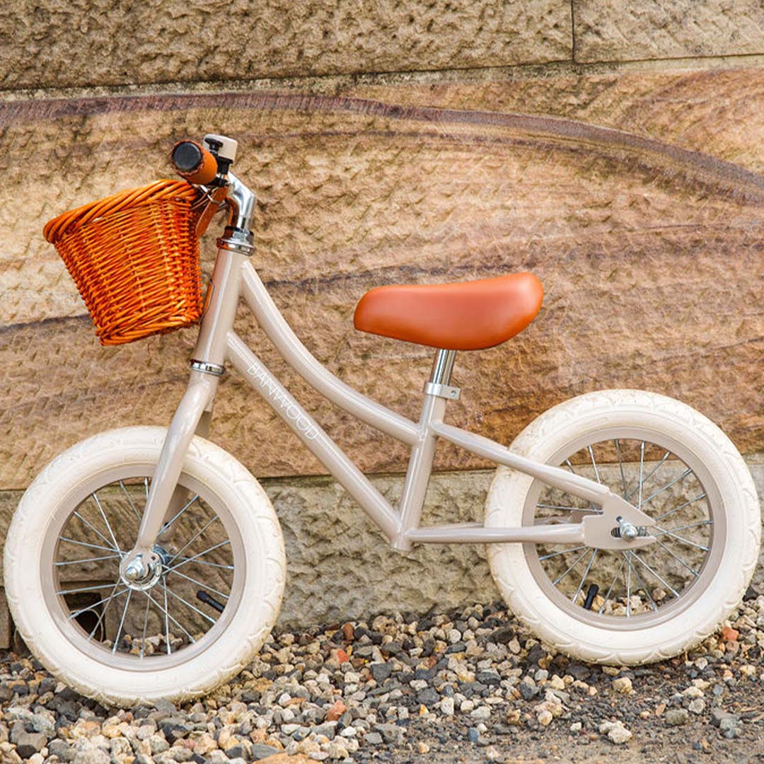 Banwood loopfiets in beige met bruin zadel en rotan mandje, staand tegen een stenen muur.