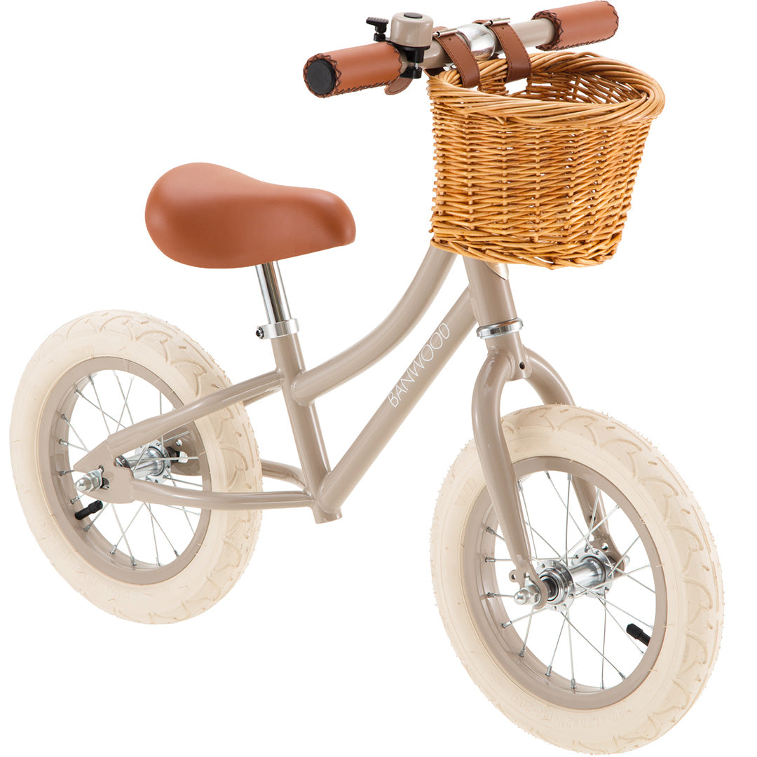 Banwood loopfiets in beige met bruin zadel en rotan mandje, schuin van voren gefotografeerd.