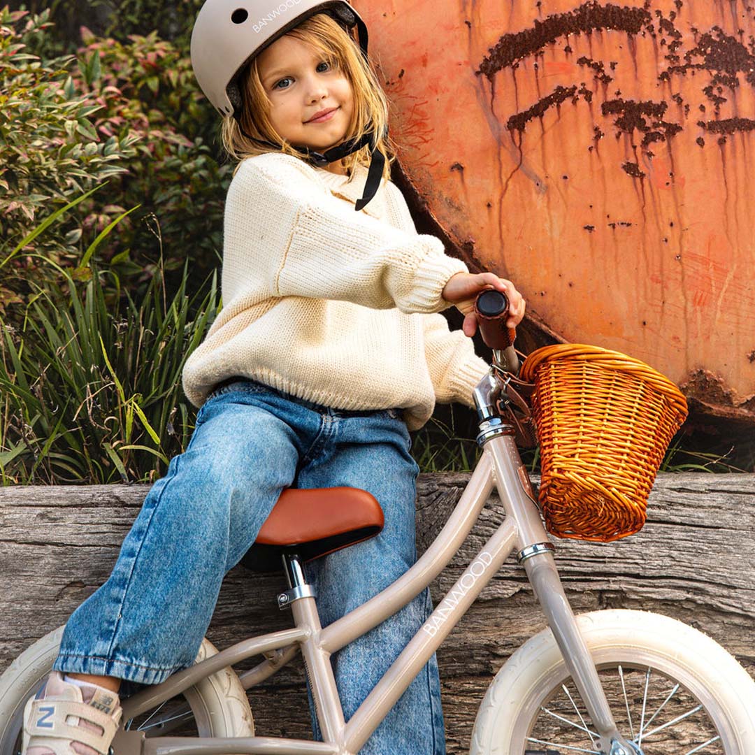 Een kind met helm zit lachend op de Banwood loopfiets met beige frame en rotan mandje.